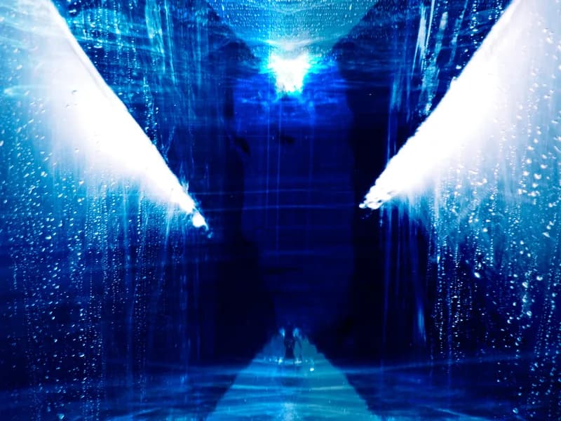 Abstract photograph in deep blue: a corridor of glassy, streaked walls converging on a bright white light, with a small dark figure on the floor below it.